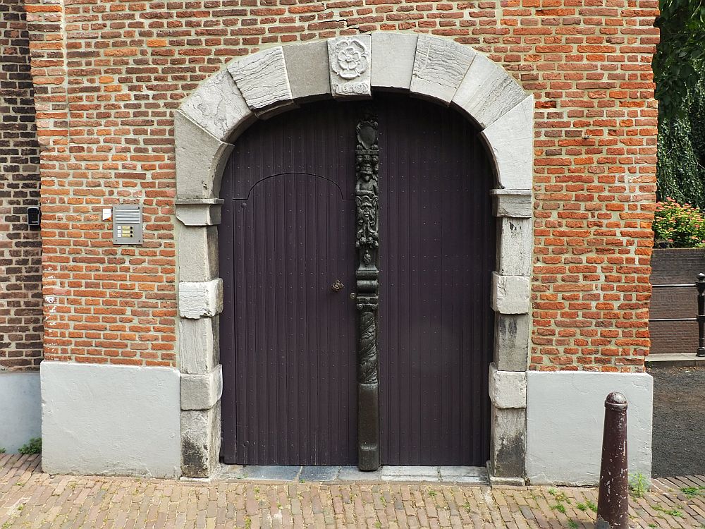 The brick building has a large archway that accommodates a double door. The door is wooden and plain, except for the vertical element down the middle, which is ornately carved. the archway is edged with stone, and the middle one above the door has a simple flower carved int oit.