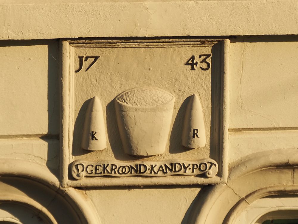 A simple plaque, rectangular. In the top left is "17" and the top right says "43". In the middle is what looks like a bucket filled with something that I presume is sugar. On either side of the bucket is a conical shape, which I think is the shape that sugar was sold in back then. The left-hand sugar cone says "K" and the right-hand one says "R". At the bottom it reads "Gekroond Kandy Pot"