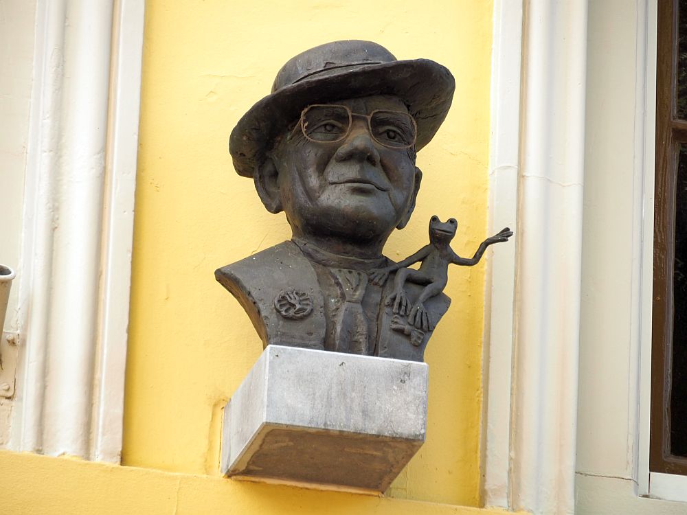 A bust of a man wearing a tie, glasses and a hat at an angle on his head. On his shoulder, a frog sits, its legs hanging down the man's shoulder and one foot/hand? waving in the air.