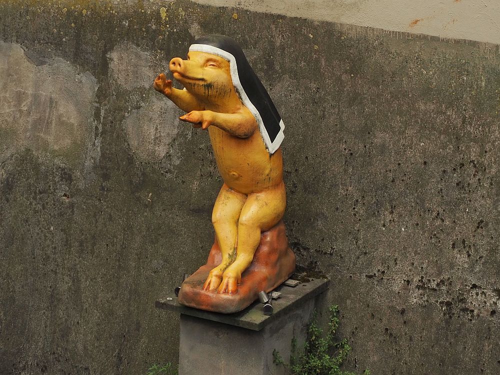 A pig sits up human style, with its front feet pointing forward. It has a nun's veil on its head, black with a white edge. Otherwise, it is naked.