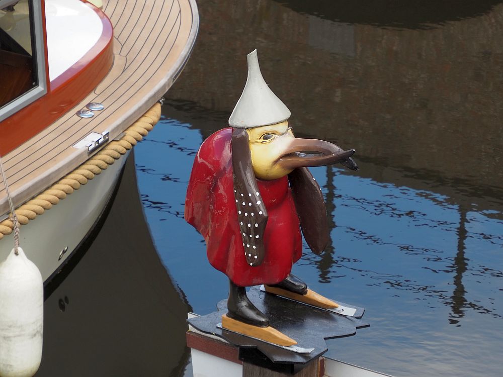 Another strange figure on a pedestal. This one is a bird, with a long beak. IT wears an inverted white funnel on its head and a red robe. It has ears or hair - brown with white polka dots - hanging down on both sides of its head. It stands on ice skates.