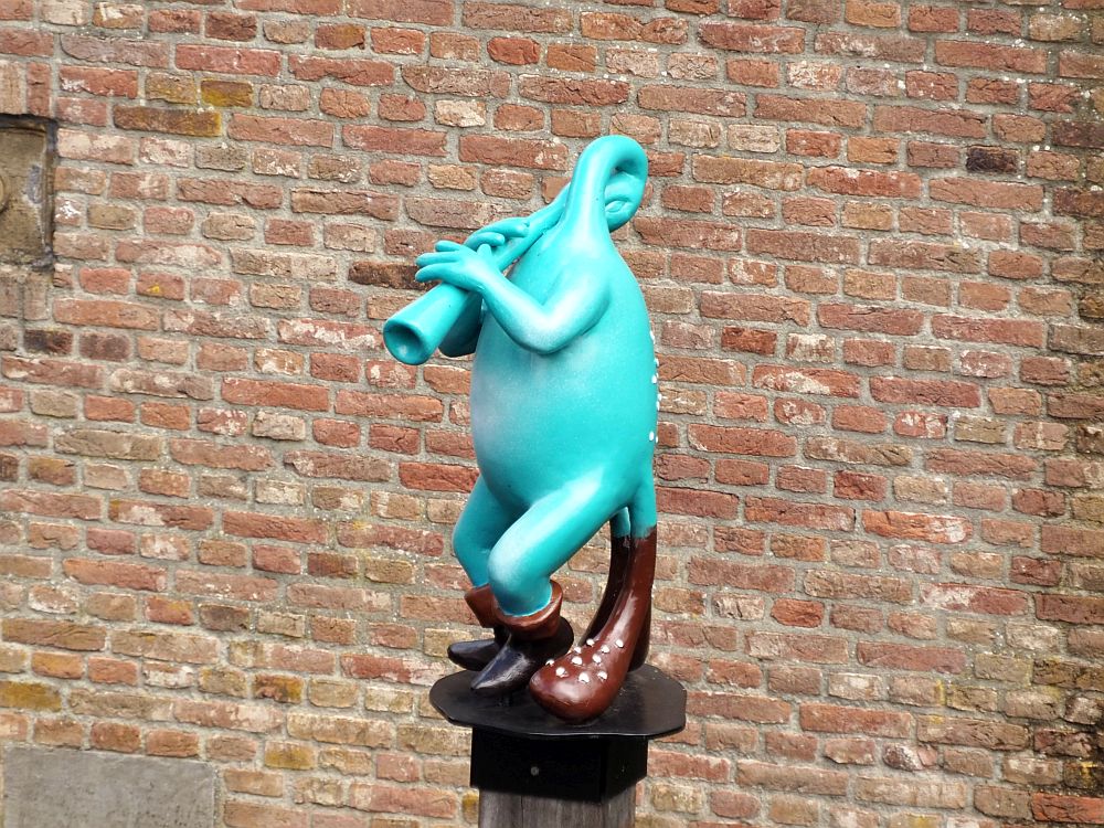 Standing on a pillar is a strange creature. It is blue and wears short dark boots on its two feet, but also seems to have two tails, which are brown with white polka dots. Instead of a head, its neck curls and takes the shape of a horn. Its arms hold the horn extending it out from its shoulder. Its hands hold the horn as if it's about to play it like a trumpet. 