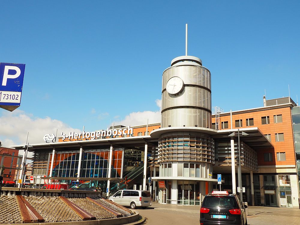 The building has a glass front and the words 's-Hertogenbosch in large letters above the front. To the right-hand side of the buiding is a large metallic cylindrical tower with a large clock on it.