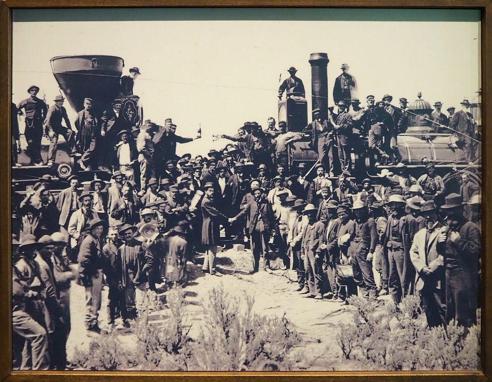 On the left and the right are two steam locomotives, pointed at each other. A crown stands in front of and on top of the locomotives, all white men, almost all facing the camera. In the middle, between the two engines, two men shake hands, also while looking a tthe camera.