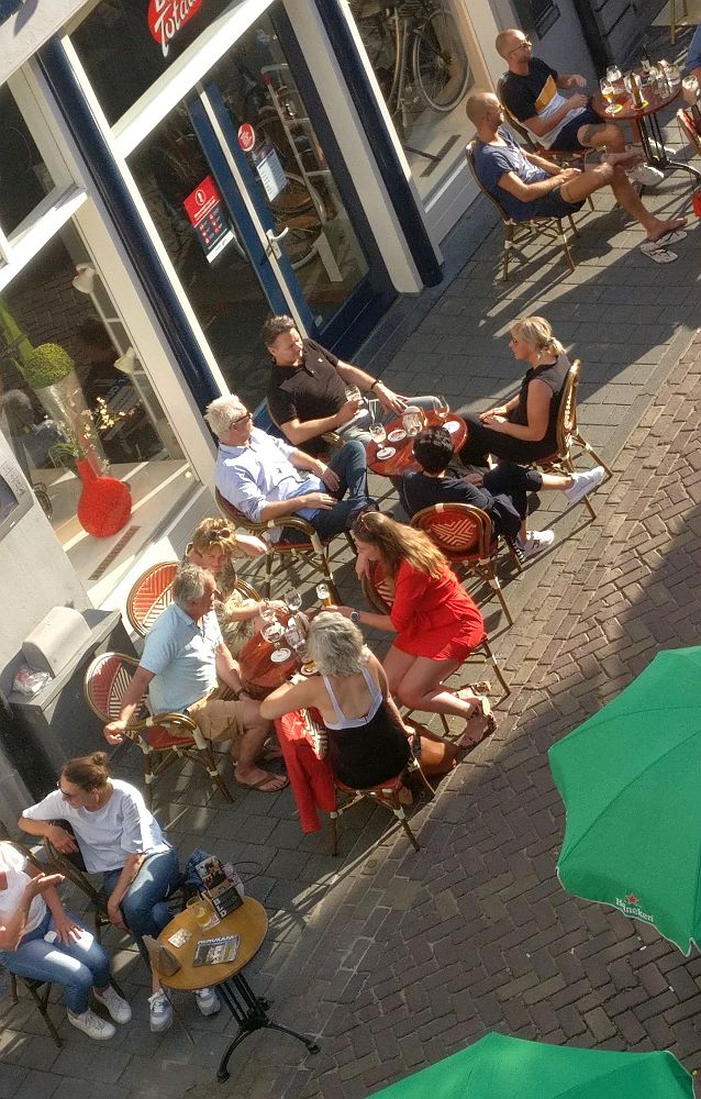 Groups of people sit outside on the sidwalk on chairs around small tables  where drinks stand. I don't know if these are family groups, but even if they are, the tables are too close together for safe distancing.