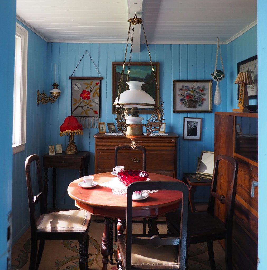 The walls of the small room are blue-painted wood. In the middle is a round wooden table with four chairs around it. A large cupboard is against the right-hand wall: it looks to be a 20th century style, perhaps 20s or 30s. An older chest is agains the far wall, and the left-hand wall has a window. A small table in the corner holds a lamp with a red lampshade. On the far wall a painting hangs above the chest with two more on either side (embroidery, possibly). A white glass lamp hangs above the center of the table, which is set with teacups.