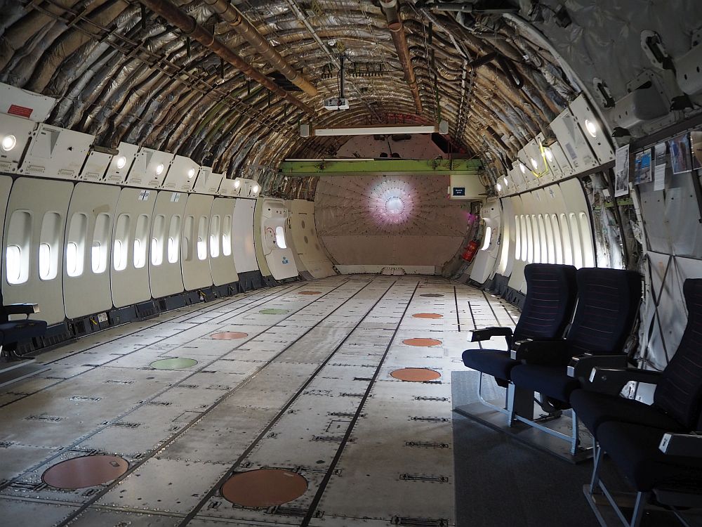 The inside is stripped down to the insulation, which is visible between the struts of the rounded ceiling. The windows still have their plastic panels that we are familiar with from fying, but the panels end at about two meters above the floor. A few airplane chairs have been placed near one wall. The floor is flat but unfinished. 