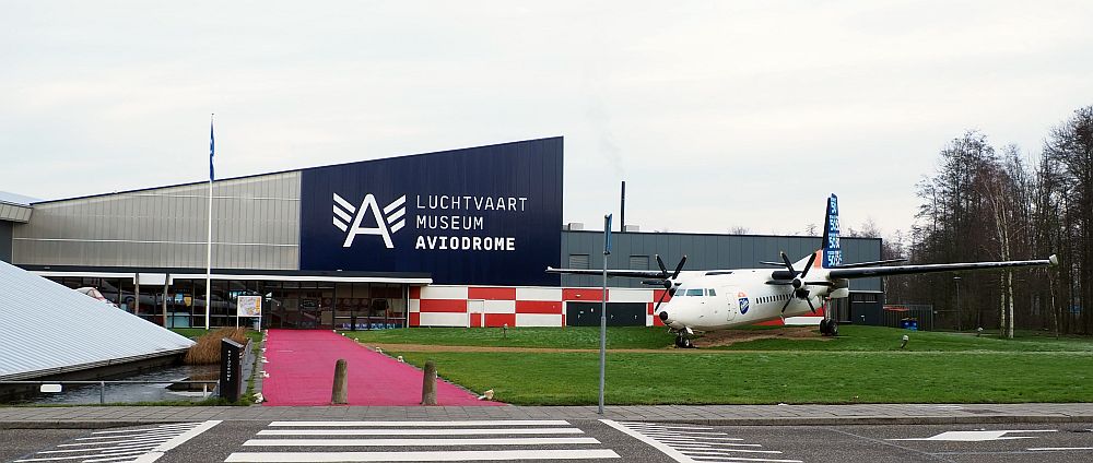 The building is metal sided, with a roof that slants up from left to right and then has another section visible behind. A large letter A logo with three stripes on either side of it is painted in white on blue above the entrance, next to "Luchtvaart Museum Aviodrome." A propeller plane is parked in front of the building.