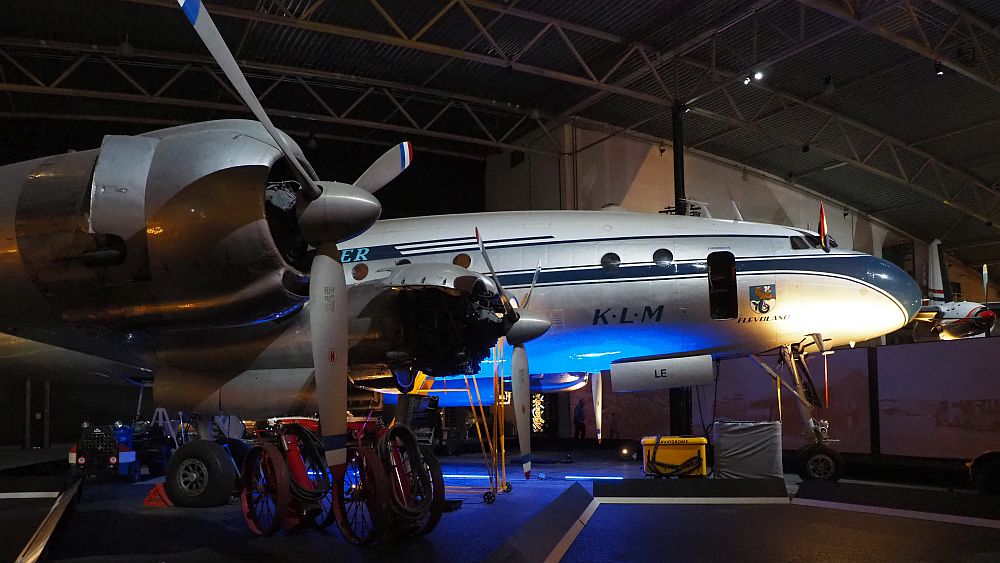 The plane is seen from straight from the side. The wing that is visible has two propeller engines sticking out in front. The body of the plane is grey with dark blue horizontal stripes. On the side it says KLM and a Dutch flag sticks out from the cockpit.