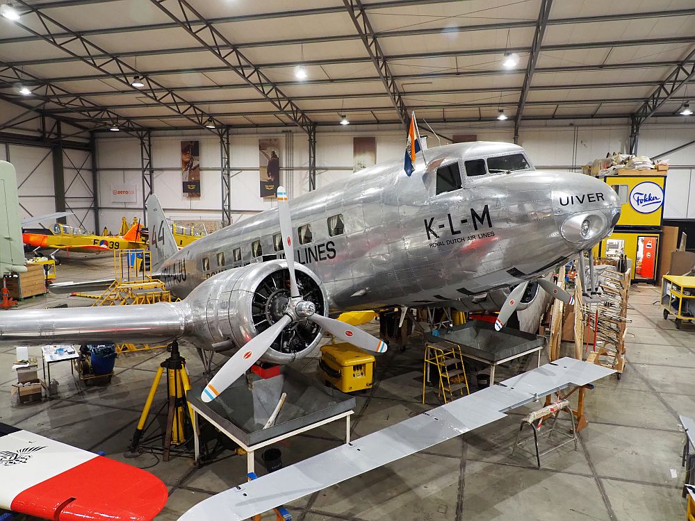 T2 Hangar at Aviodrome Lelystad is a much more brightly-lit room than in the rest of the pictures. A shiny grey metallic plane with two propellers. It has KLM and Uiver painted on the nose. It slants upward to the right, and has an assortment of tables underneath it. Its windows are square and have curtains neatly pinned back (other than the cockpit windows, which have no curtains.