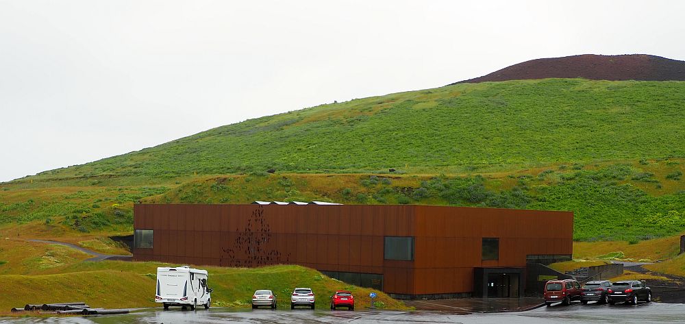 The building is a simple, rust-colored block, partly built into the side of the hill/volcano that rises behind it. A few cars are parked in front of the building. The hill behind is covered in grass, except for a very dark brown top.