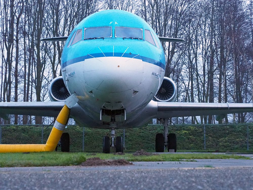 A modern (ish) jet plane seen from directly in front: the nose mostly fills the picture, and only part of the wings are visible, extending out below the belly of the plane. The engines are visible as two circles. The plane is painted in KLM colors: blue above white.