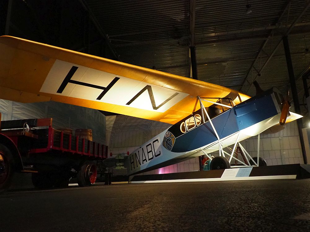 The body of the plane is boxy and mostly blue, with one propeller on the front and the letters HNABC painted on the side. The wing (only one is visible in the photo) is yellow, with the letters HN painted on the underside. Three small windows in the body are where the passengers sat. 