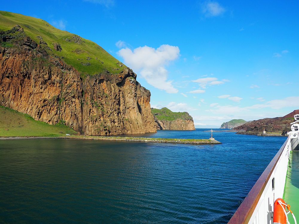 Looking down the side of the ferry, which is along the right side of the photo. Ahead is a view of blue water and the narrow channel the ship passes through. A high brown cliff on the left with a grassy top. More cliffs of the same type further down the channel.