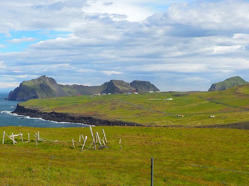 A view across the island of Heimaey, Iceland, in the Westman Islands. Flat green grass in the foreground, with cliffs on the left dropping into the sea. In the background, a ridge of mountain.