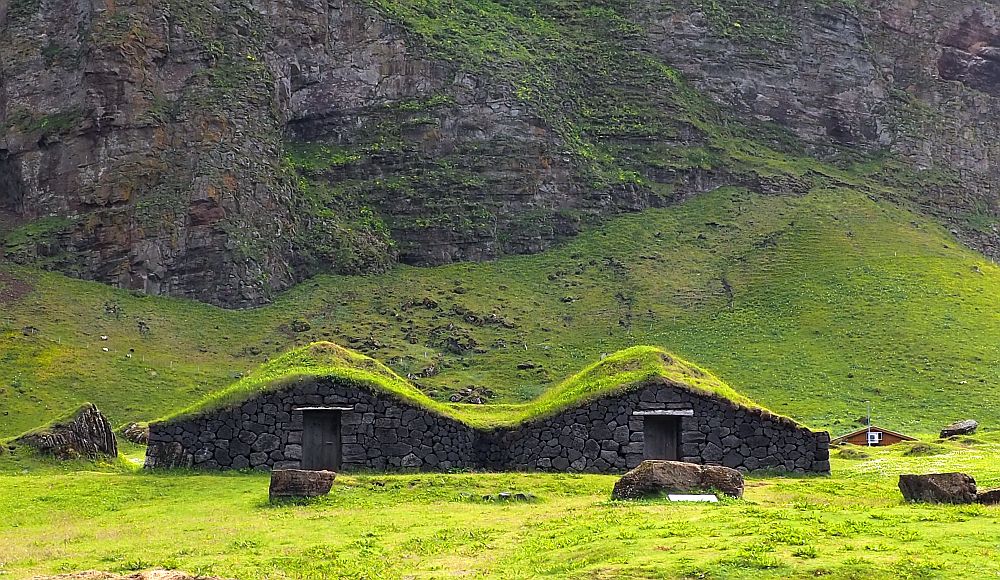 Seen from the front, each of the two parts has a peaked roof covered in grass. The center of each partk has a single doorway and no windows. The fronts are stone. Behind it is a steep cliff.