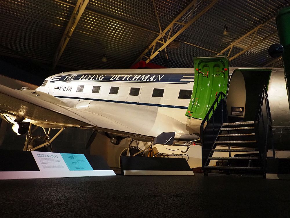 A more modern-looking airplane slants up to the left. It is painted white with a blue stripe along the length of it, in which the words "THE FLYING DUTCHMEN" are painted in white. The windows are rectangular. The door is open and a short flight of stairs leads up to it.