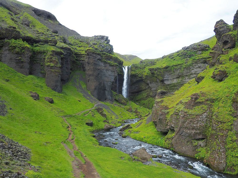 View down the canyon: cliffs on both sides of exposed rock, with grass growing on the less steep surfaces. A river snakes through and the waterfall is visible in the center of the picture: a long narrow shape falling off a cliff.