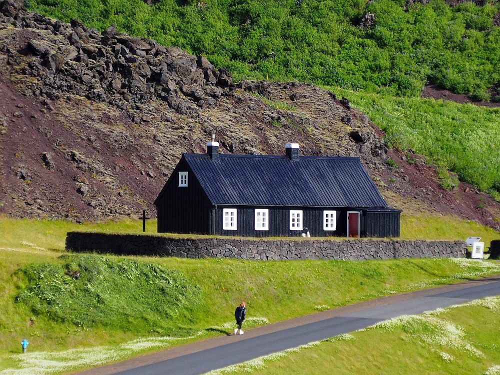 A simple wooden cottage with black shingle roof and sides and white widows. It's a simple rectangle with two chimneys. A low stone wall around it topped with grass. 