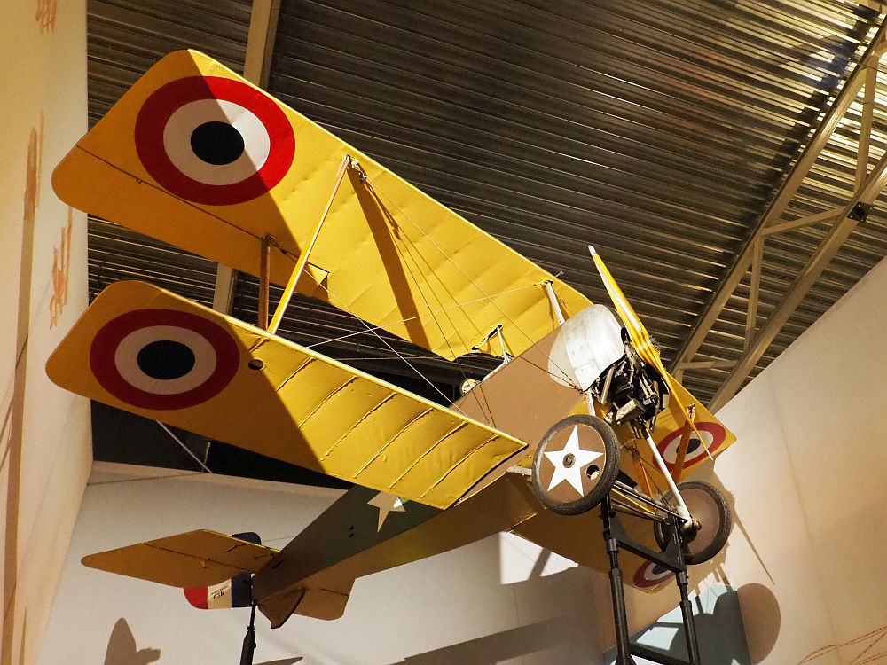 A bi-wing plane, seen from below (It hangs from a corrugated metal ceiling. It has one propeller in front and the wings are painted yellow with a bright round spot at the end of both top and bottom wings: black in the center, then a ring of white, then a ring of red. The body is brown, and the wheels just behind the propeller have white stars painted on them.