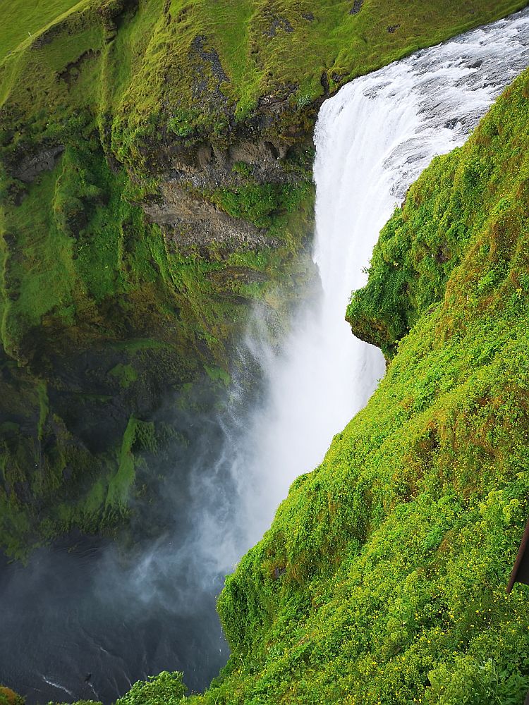 The waterfall plunges down from the top right of the photo toward the bottom left. The water mists outward as well as falling downward. The ground on either side of it is green with low growth.