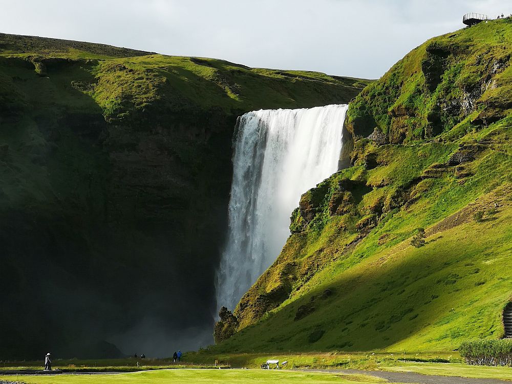 Skogafoss falls off a horizontal shelf in one straight waterfall. On either side of that shelf: green steep hills. A  person at the bottom left gives a sense of the size of this waterfall. 