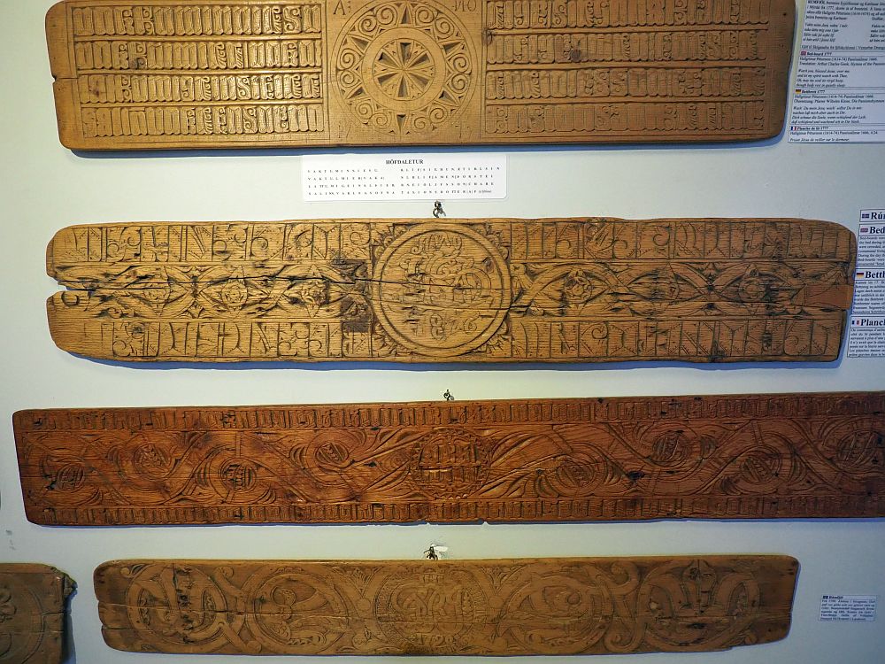 Four bedboards in this photo. Each is more or less rectangualr, but somewhat irregular at the ends. Each has intricate carvings with small primitive images and also lettering.