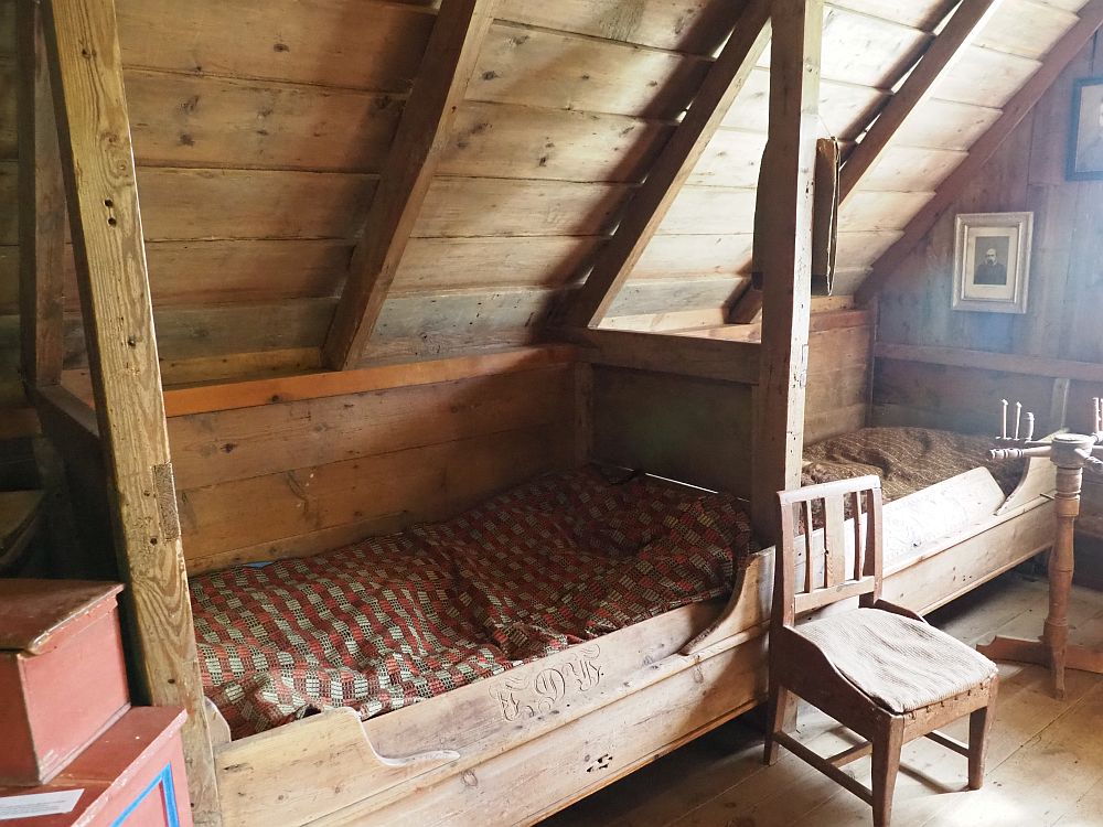 The ceiling leans inward under the roof. It is made of wood. The two beds, end-to-end, also wood, are built into the wall and attached to each other. Each has a bedboard along the outside edge with a simple carving of lettering on it. Each has bedding that looks thin and bumpy with a cloth or sheets on top. 