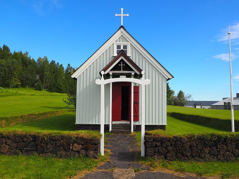 Seen from the front, the church is simple and white, with a red door, a white entrance gate, and a simple white cross on the roof instead of a steeple.