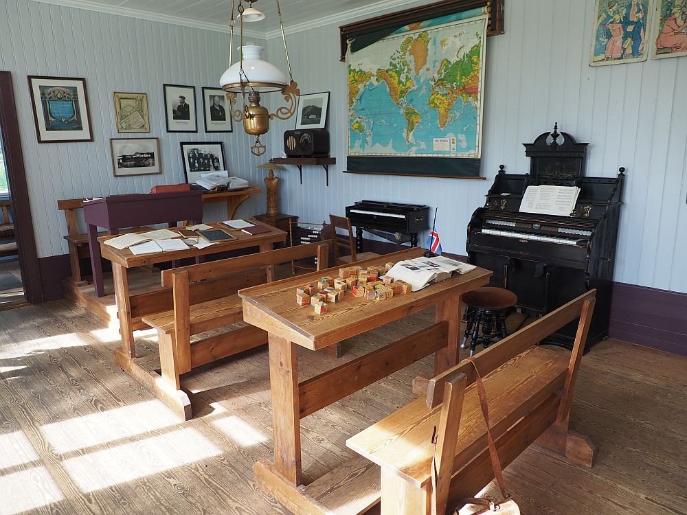 Only two wooden desks with benches fixed to them. Each would sit perhaps three children. The teachers desk is beyond them and set on a small platform. On the wall to the side is a piano and what may be an organ. A world map hangs on the wall. 