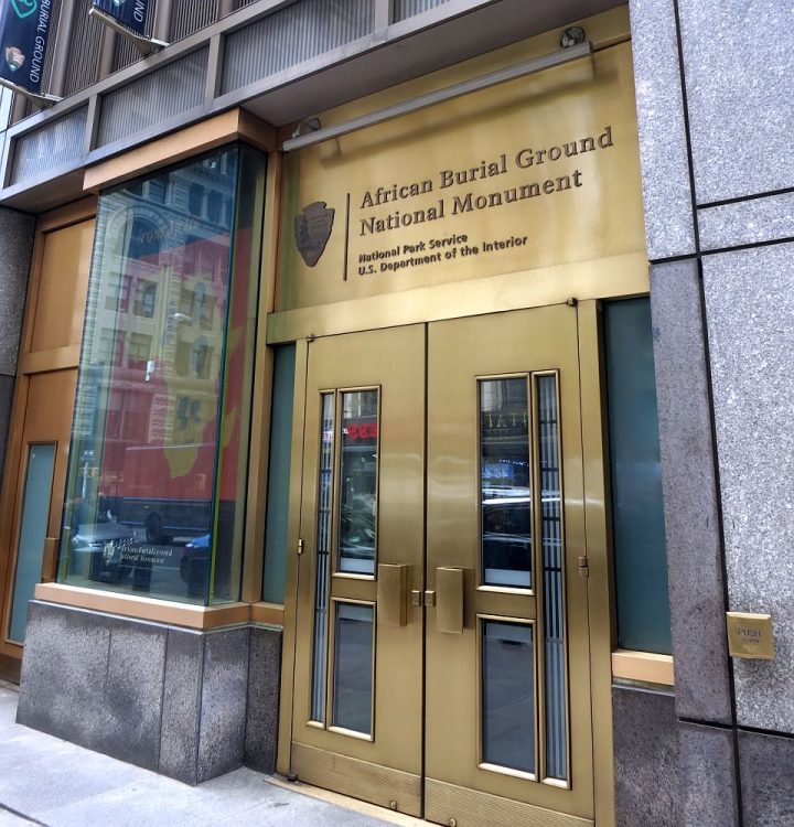 View of the entrance to the African Burial Ground: a double door that's a bronze color with glass panes down the middles, with a bronze panel above it saying "African Burial Ground National Monument, National Park Service, US Department of the Interior.