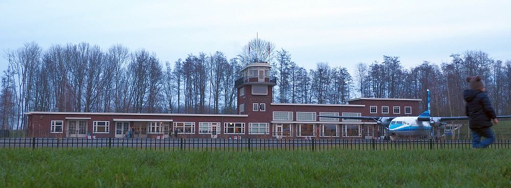 A long, low, brick building, mostly just one story, though on the right-hand end it's two stories. The control tower is in about the center and is about four stories high. It's al very simple, with white-edge windows, and a small balcony around the top floor of the control tower. In front of the building, to the right, is an airplane.