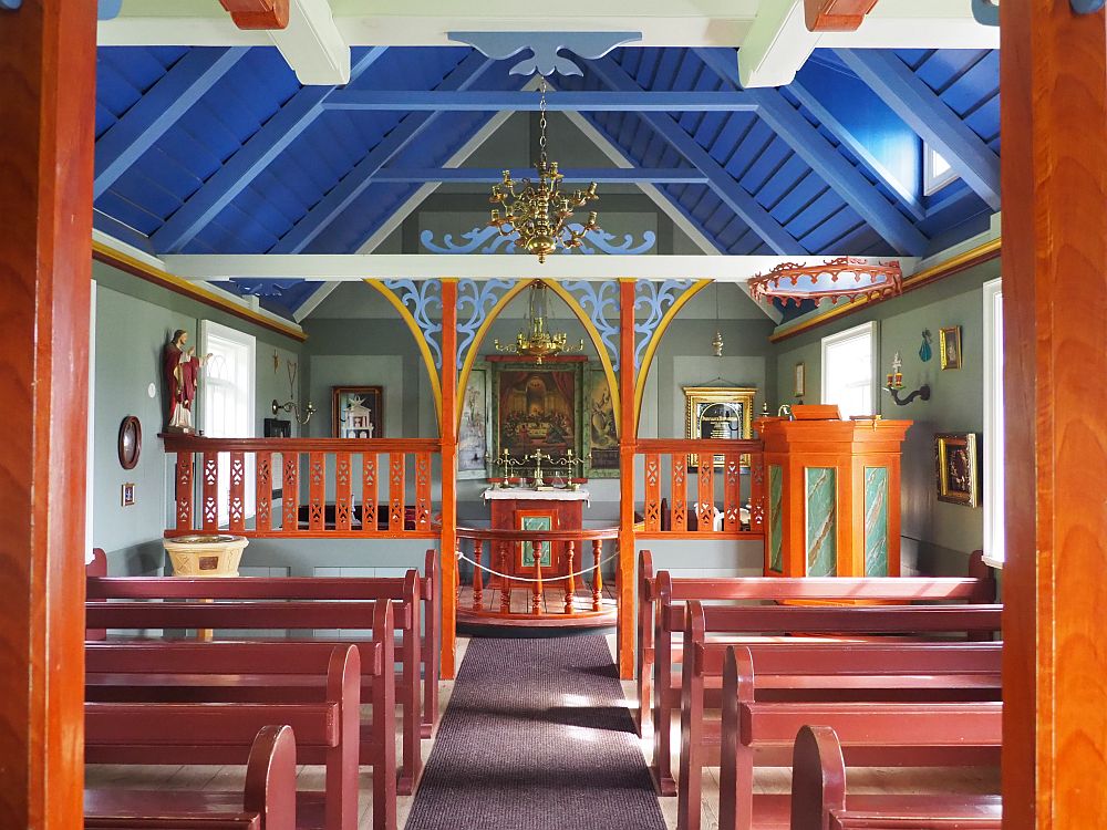 Looking down the aisle, pews on either side, painted reddish pink, only about five rows on either side of the aisle. The  altar, straight ahead, has a painting above it and a bronze chandelier above it. Two more paintings are on either side of the altar and a statue of Jesus (?) is on the left-hand wall. The pulpit is before the altar on the right: wood but with a marbled inset on each of 4 or five visible sides. The walls are light green and the ceiling is a bright blue.