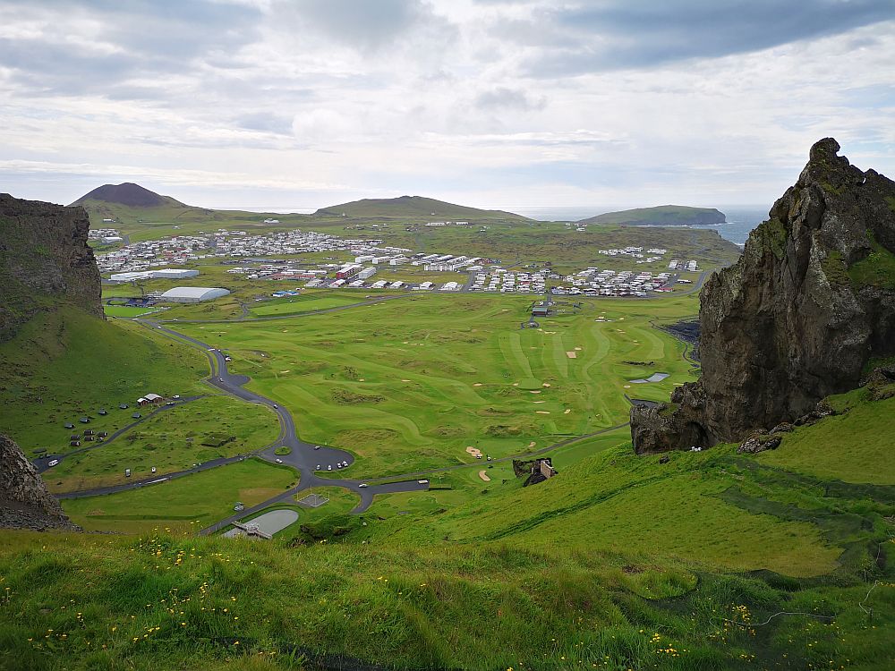 With craggy rocks on the left and right, the middle of the phot is green. Below, looking tiny, is the parking lot, and beyond that is more green: a golf course. Beyond that are some neatly lined-up, low builidngs. 