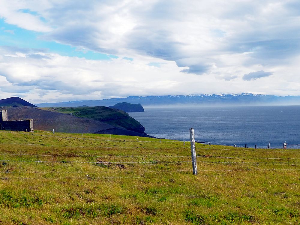 A view across a grassy field with a few wooden fenceposts. Beyond the field is the sea, and beyond that is a mountain range with white peaks. 