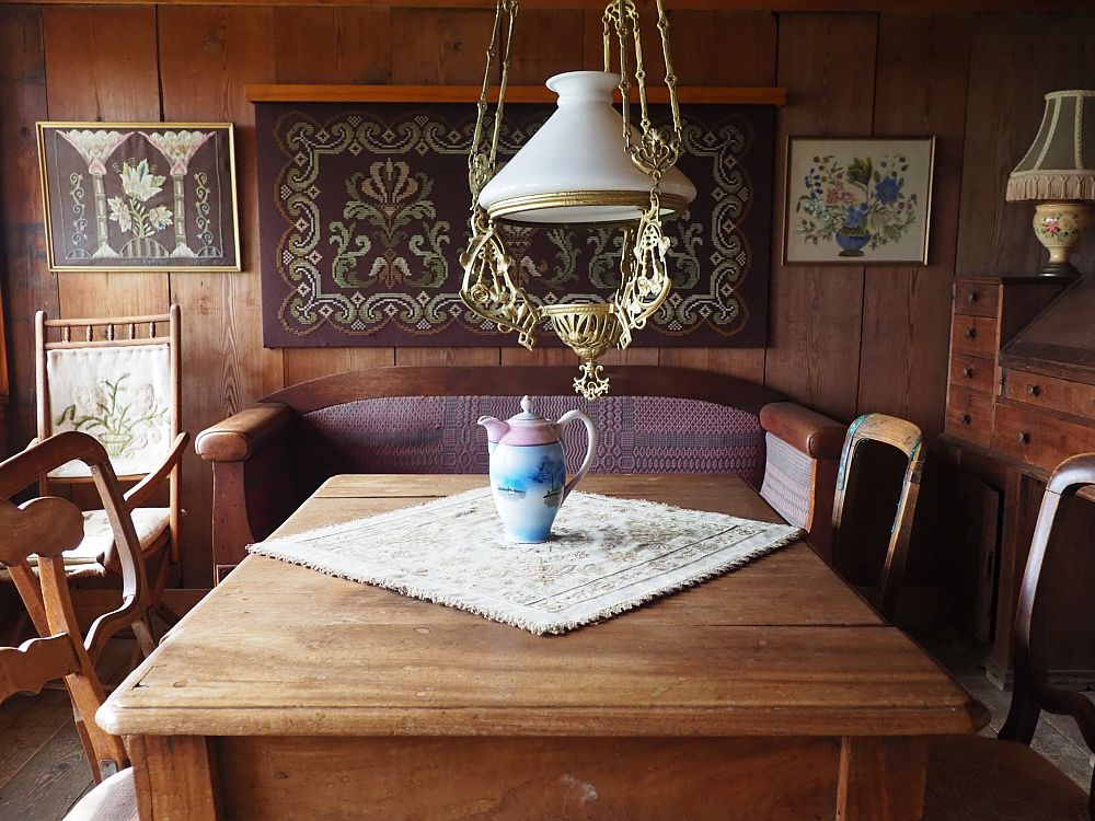 The rooms is mostly filled with a rectangular wooden table and the four chairs around it. A white glass lamp hangs above it. Beyond is a sofa against the far wall. Above the sofa is what looks to be a decorative rug with embroidery on either side of it.