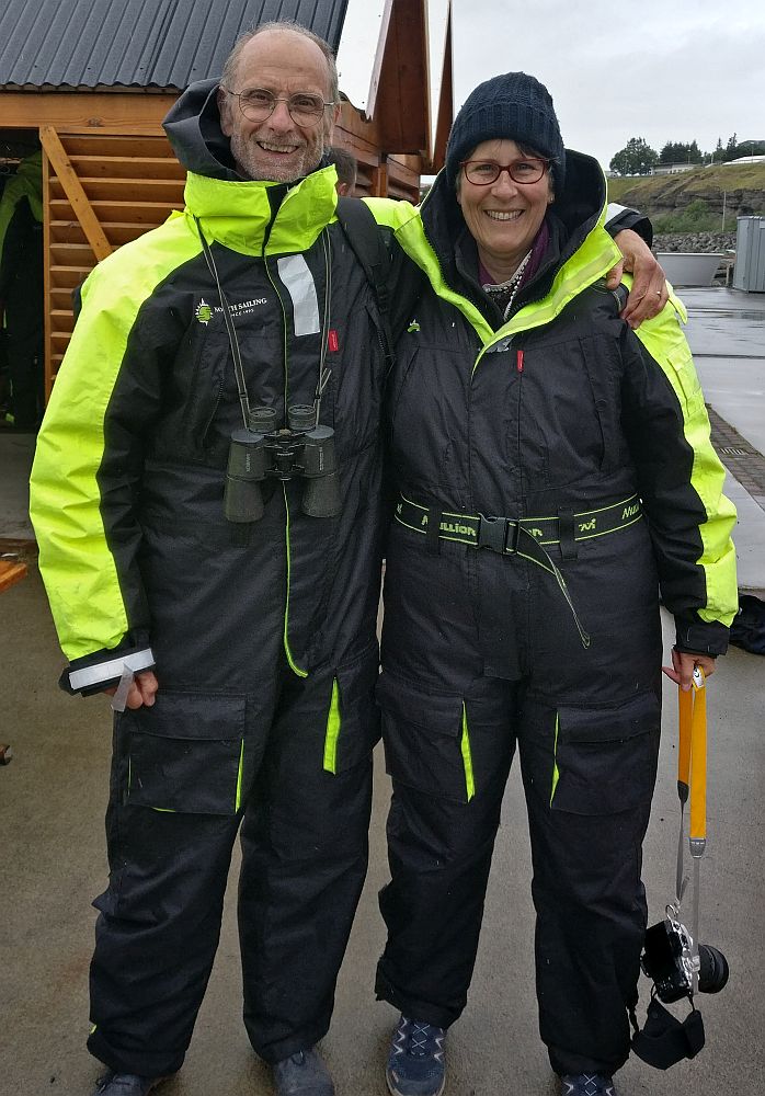 Albert and I pose facing the camera, arms around each other's shoulders. Both wearing a one-piece coverall, black, but with bright yellow along the tops of the sleeves and around the collar. I have a camera dangling from my hand and a warm cap on my head. Albert has a pair of binoculars hanging from his neck.