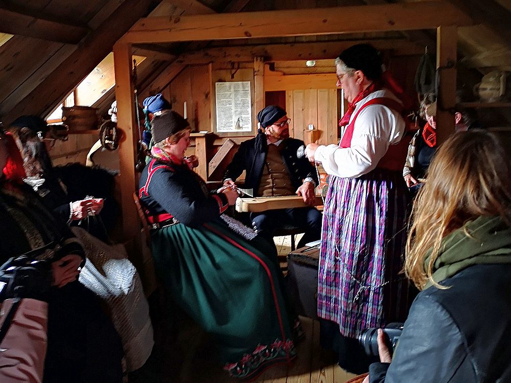 A crowded room. the ceiling slants inward with a window in the slanted ceiling to the right. In the center back, a man sits with the instrument on his lap. He wears an old-fashioned black jacket and a black hat. ON the left and right are women in long skirts, wearing shirts and embroidered vests and black hats. 