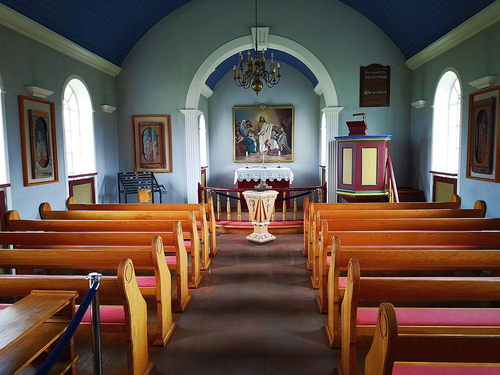 Looking straight down the aisle toward the altar. Pews on either side: wood with red seats. At the end of the room is an archway with a railing dividing it from the main room. A religious painting on the far wall, over a lace-covered altar. The pulpit is before the archway to the right, and painted simply: dark red and yellow. The ceiling is arched and dark sky blue. The walls are light blue with white trim.