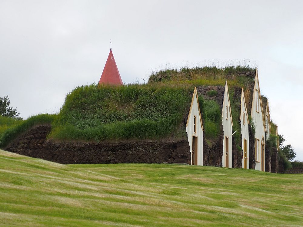 Seen from a side view, some of Glaumbaer's front gables are visible: white with a mustard-yellow trim. They vary in size, but none is more than two storeys. All are covered with grassy roofs. A red church steeple peeks up behind the turf farmhouse. 