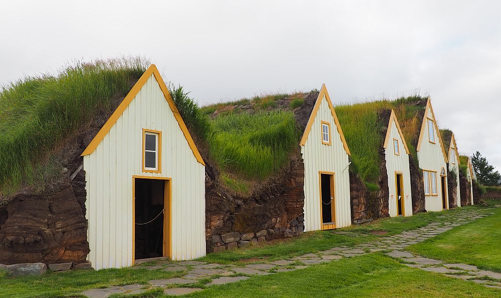 The gables look like how a child might draw a house: door in the center, one window above the door. Each is painted off-white with yellow trim along the roof line and around windows and doors. Between the gables is turf covered with grass. 