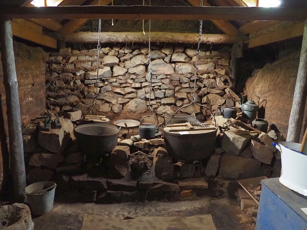 A wall of rocks lines the back wall of the room. In front of that is a shelf of rocks. Several pots sit on the rocks or hang from chains from the ceiling. A kettle and some smaller pots stand on the rocks to the right. The walls on the left and right are bare turf and the floor is dirt. The light comes from two skylights, one on each side in the roof above the hearth.