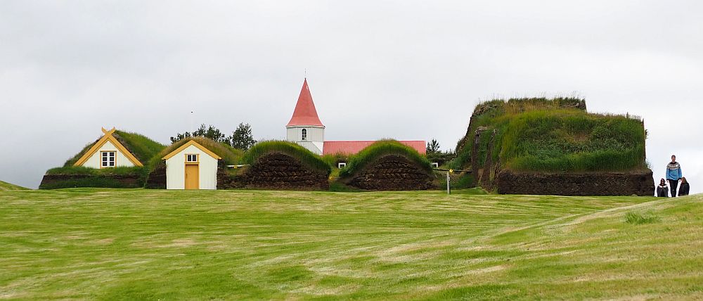 From the side only 2 gables are visible to the left. Most of the structure looks like a bunch of grassy lumps. The steeple of the church can be seen behind the house: it is pointy and red.