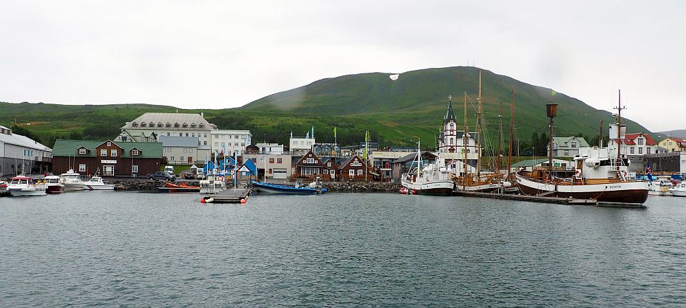 Husavik whale watching leaves from this harbor. Cluster of buildings along the water's edge, several large oak boats moored along a pier on the right. A mountain behind the village with a bit of snow on top. Grey water in the foreground, white sky.