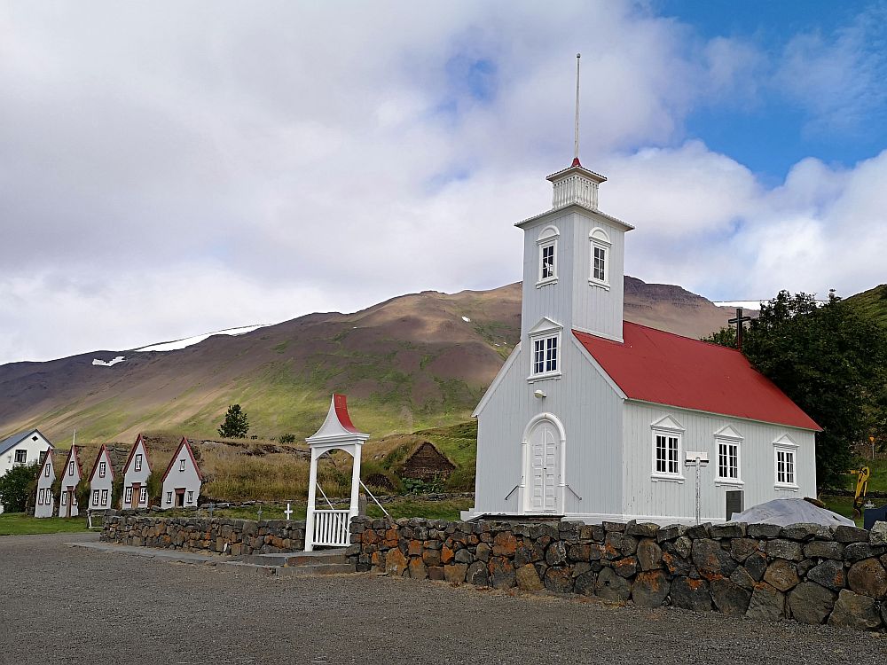 In the forground, the church is white and a simple rectangle, with a door in the short end and a window right above the door, a steeple (perhaps one additional storey tall) above that. The long side of the rectangle that is visible has 3 evenly-spaced windows. The roof is reddish. Beyond the church are the 5 gable fronts of Laufas turf house. Behind them is a mountain.