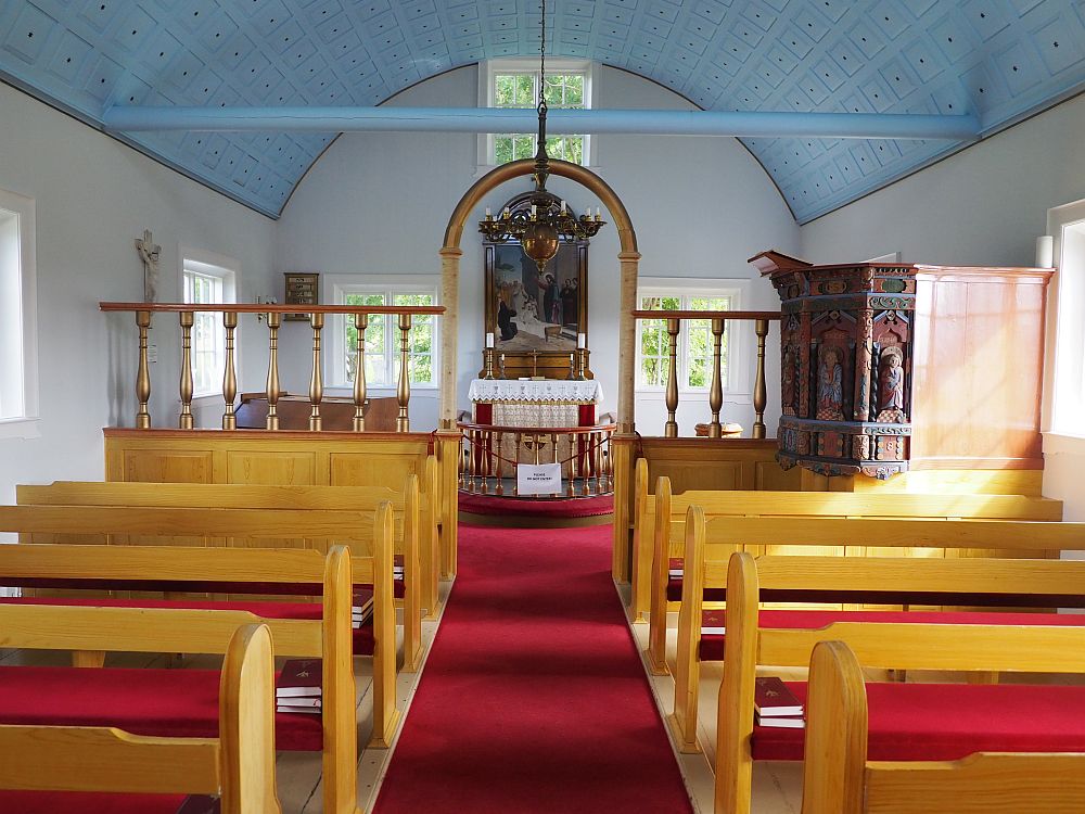 Looking down the aisle of the church. Pews on either side, wood with red seats. A railing dividing the pews from the altar. The altar has a large painting above it. On the right, before the railing, is the old pulpit, carved with saints. The walls are clean white and the ceiling is arched and painted light blue.