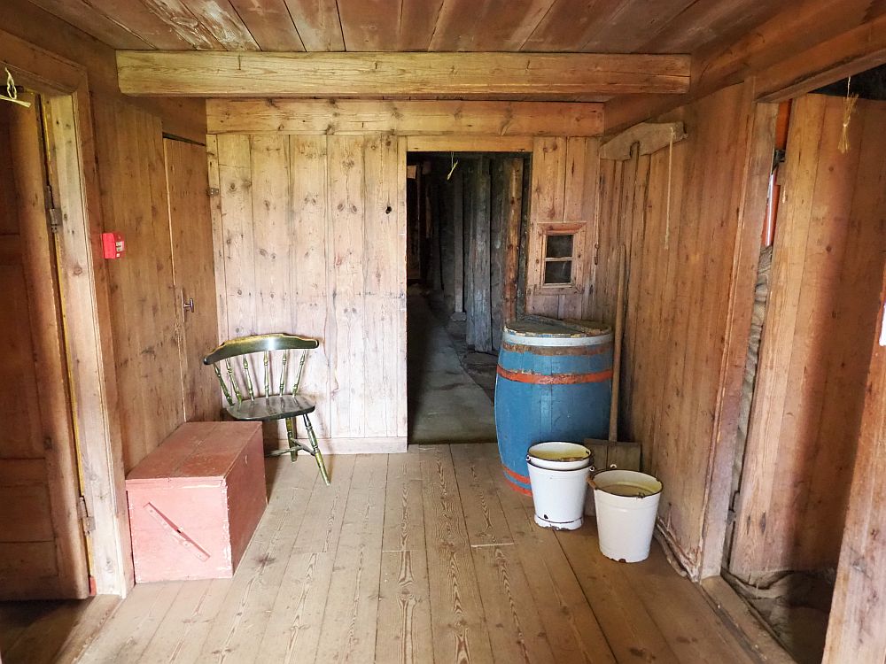 A square space, with boards for walls, floor and ceiling. A door to the left and a door to the right. Straight ahead, another door in the back wall, off-center. Through that one, a bit of a hallway is visible with large poles lining it. A spindle-back chair in the left back corner, with a small trunk between it and the left-hand door. A large wooden barrel in the back right corner, with several metal buckets next to it.