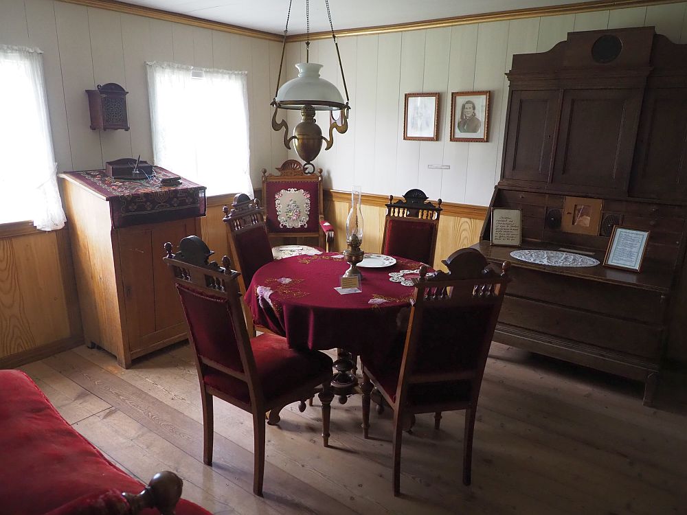 The floor is wood boards, and the bottom half of the walls are panelled. Above the paneling the walls also appear to be wood boards, but painted off-white. On the left back wall are two windows, light coming through white curtains. Between the curtains is a writing table with a slanted top. In the far corner is an upholstered red chair. On the right back wall two portraits hang and then next to them is a large cupboard, perhaps a secretary. In the middle of the room is a round table covered in heavy red cloth with an oil lamp in the center. Around it are four upright chairs upholstered in red. On the near left wall (the photo was taking from a door in the corner) a bit of a red upholstered sofa is visible.