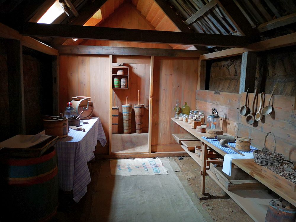 The room has a dirt floor and a peaked ceiling. Along both left and right walls are long tables lined with various kitchen tools. The walls are wood paneled to about 2/3 of the way up, but then above that the turf bricks behind the paneling is visible. Light comes in through a skylight in the roof on the left. A second room is visible through a doorway at the end of the room. There, the floor is wood and a bit higher. Butter churns can be seen standing against the far wall. 