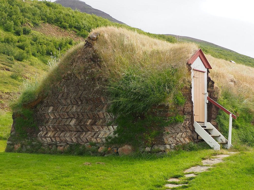 The "house" looks almost like a grass-covered hill except that the side facing the camera has visible brown turf bricks in a herringbone pattern. On the side is a door leading into a grassy turf wall. A few short steps lead up to the door.